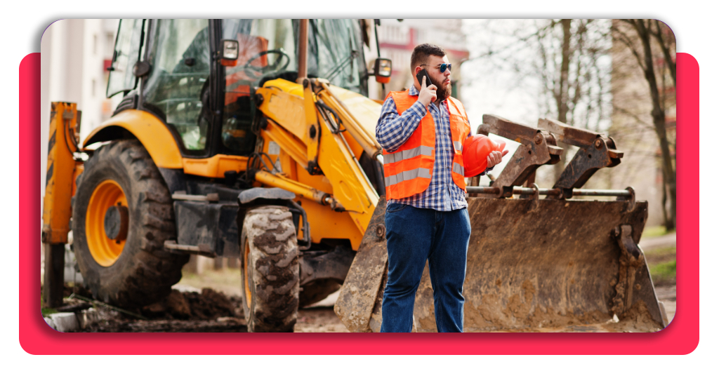 Consigue trabajo ya: curso de maquinaria de movimiento de tierras 2 Trabajador de la construcción con casco naranja frente a un tractor con teléfono móvil en mano